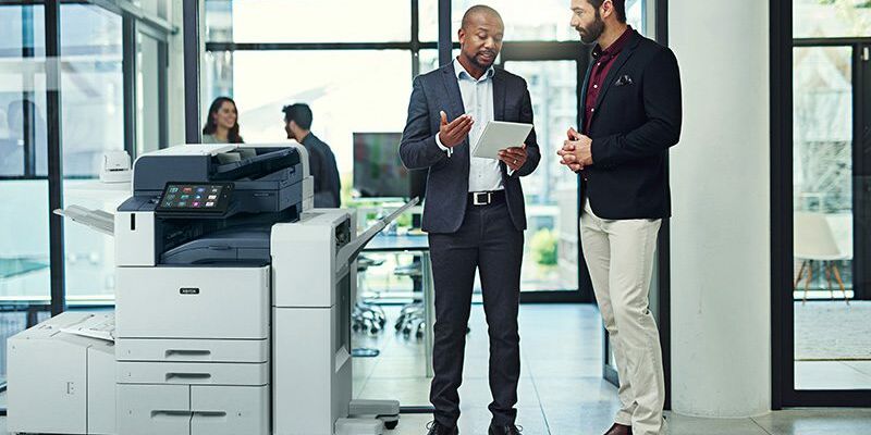 Deux hommes dans un bureau discutent sur une tablette &agrave; c&ocirc;t&eacute; d'une imprimante multifonction couleur Xerox&reg; AltaLink&reg; s&eacute;rie C8200