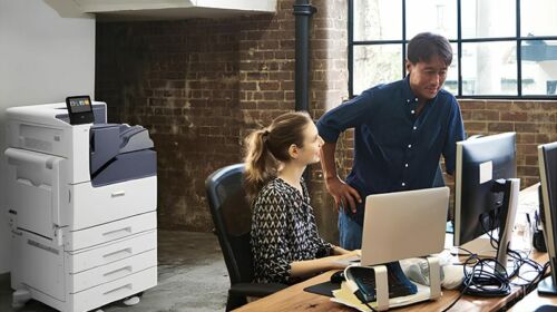 Un homme et une femme se tiennent côte à côte devant un ordinateur, concentrés sur l'écran. L'imprimante couleur Xerox® VersaLink® C7000 est en arrière-plan.