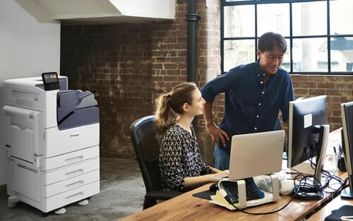 Un homme et une femme se tiennent côte à côte devant un ordinateur, concentrés sur l'écran. L'imprimante couleur Xerox® VersaLink® C7000 est en arrière-plan.