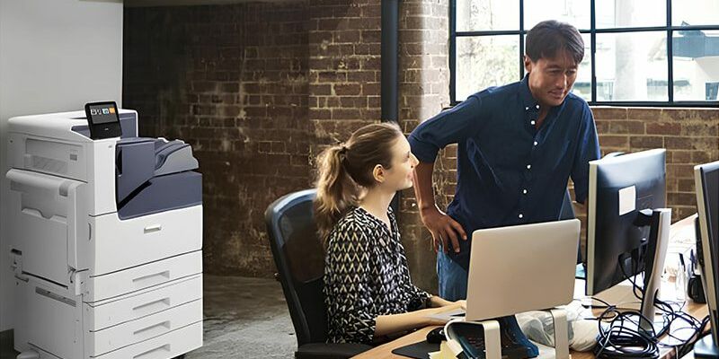 Un homme et une femme se tiennent côte à côte devant un ordinateur, concentrés sur l'écran. L'imprimante couleur Xerox® VersaLink® C7000 est en arrière-plan.